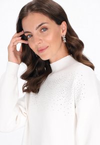 Maglione bianco con collo alto e un motivo testurizzato di piccoli strass sulla parte anteriore superiore. Il modello ha capelli mossi e marroni e indossa orecchini.