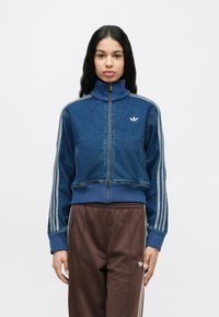 Geacă din denim albastru cu guler înalt, închidere cu fermoar și logo alb Adidas. Prezintă accente cu dungi gri pe mâneci.