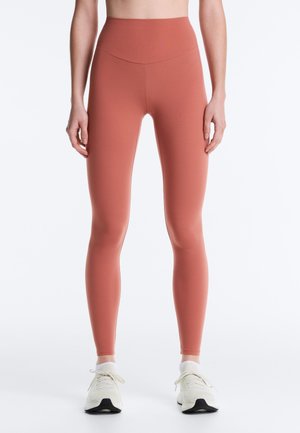 Kobieta ubrana w legginsy z wysokim stanem w kolorze rdzy oraz białe buty sportowe, stojąca na tle jednolitego białego tła.