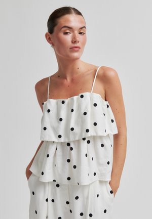 Kvinne iført hvit ermeløs lagdelt topp og bukser med svarte prikkete mønster, stående med hendene i lommene mot en ensfarget bakgrunn.