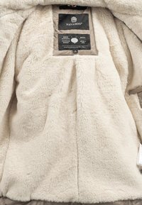 Cream faux fur interior of a jacket, featuring a black label with brand and sizing information. Smooth, soft texture throughout.