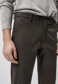 Homme portant un pantalon brun foncé et un pull gris clair, montrant sa taille et sa main détendue sur le côté contre un fond uni.