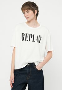 Pessoa a usar uma t-shirt branca com o texto "REPLAY" e jeans azul-escuro, posando com uma mão no bolso contra um fundo neutro.