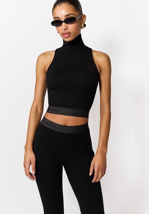 Kobieta ubrana w czarny, bezrękawnik z wysokim kołnierzem i krótki top oraz dopasowane, czarne spodnie z wysokim stanem i elastycznymi pasami, czarne okulary przeciwsłoneczne i złote kolczyki-koła.