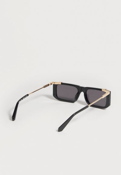 Lunettes de soleil rectangulaires noires avec branches métalliques dorées et verres teintés foncés, reposant ouvertes sur une surface blanche.