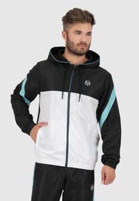 Hooded jack met een zwart-wit kleurblokontwerp, voorzien van turquoise accenten en een elastische zoom. Gemaakt van een lichtgewicht materiaal.