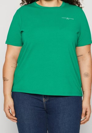 Persoon die een groen Tommy Hilfiger T-shirt met korte mouwen en donkerblauwe spijkerbroek draagt, met zichtbare armtatoeages en krullend haar.