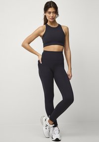 Sort sportsbra og højtaljede leggings lavet af glat stof, med et tætsiddende design og sømløs finish. Båret med hvide træningssko.