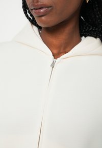 Hoodie sa zatvaračem u mekom kremastom materijalu, s metalnim zatvaračem srebrne boje i širokom, mekanom kapuljačom. Blizak prikaz ovratnika i detalja teksture.