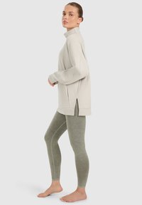 Femme debout pieds nus, portant un pull oversize beige clair avec des fentes latérales et des leggings ajustés vert olive sur un fond uni.