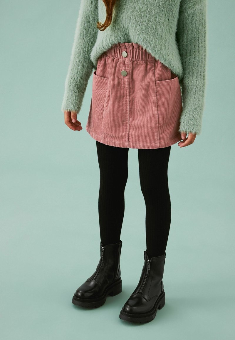 Fille portant un pull duveteux vert clair, une jupe en velours côtelé rose avec des boutons et des poches, des leggings noirs et des bottes noires zippées sur un fond uni.