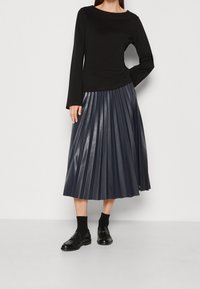 Haut noir à manches longues associé à une jupe en cuir plissée bleu marine longueur genoux et des chaussures lacées noires, présentant des motifs texturés et des finitions lisses.