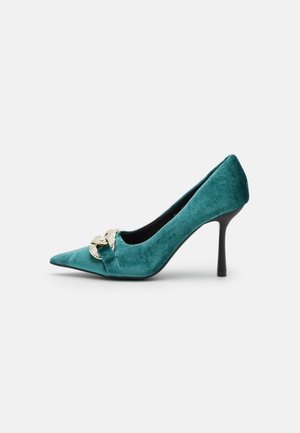 Buty na wysokim obcasie z zielonym welurem, z ostrym noskiem, czarnym obcasem typu szpilka i ozdobnym złotym łańcuszkiem z przodu.