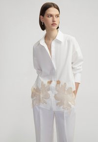 Camisa blanca de gran tamaño con cuello puntiagudo y mangas largas, con bordado floral beige cerca del dobladillo. Textura de tela suave.