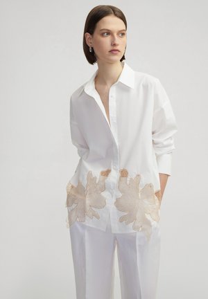 Camisa blanca de gran tamaño con cuello puntiagudo y mangas largas, con bordado floral beige cerca del dobladillo. Textura de tela suave.