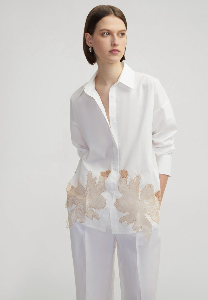 Camisa blanca de gran tamaño con cuello puntiagudo y mangas largas, con bordado floral beige cerca del dobladillo. Textura de tela suave.