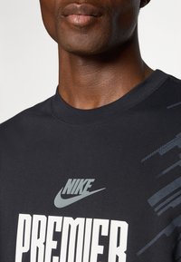 Sort Nike t-shirt med hvid "PREMIER" tryk og et gråt logo. Glat stof med rund hals og subtile geometriske detaljer.