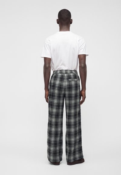 Homme debout de dos, portant un t-shirt blanc uni et un pantalon ample à carreaux noir et blanc, sur un fond gris uni.