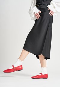 Zapatos planos de piel roja con correa y hebilla circular, llevados con calcetines tobilleros blancos con volantes. Textura suave, puntera afilada, diseño minimalista.