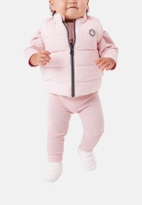 Ljusrosa vadderad väst med hög krage, dragkedja och marinblå detaljer, kombinerad med matchande rosa leggings och vita skor.