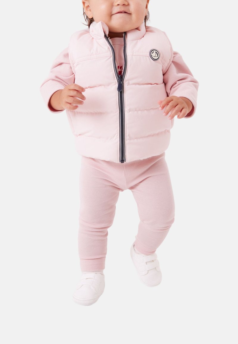 Ljusrosa vadderad väst med hög krage, dragkedja och marinblå detaljer, kombinerad med matchande rosa leggings och vita skor.