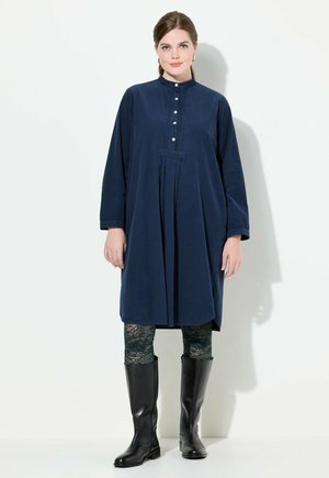 Marineblauwe jurk met lange mouwen, knoopsluiting aan de voorkant, geplooid ontwerp, gecombineerd met groene kantpatroonlegging en hoge zwarte laarzen.