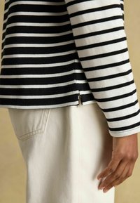 Mustriga must-valge pika varrukaga topp, millel on külje lõhik ja mis on kantud heledate pükstega, millel on nähtav tagakoti õmblus.