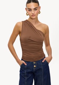 Haut marron à une épaule en tissu extensible avec une texture froncée. Associé à un pantalon en denim taille haute avec fermeture à bouton et poches.