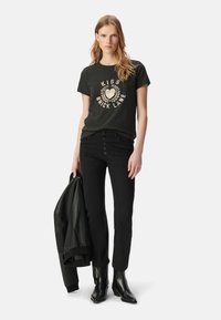 Jeans negros con cierre de cinco botones, combinados con una camiseta verde oscura con un gráfico crema. El modelo sostiene una chaqueta de cuero negra.