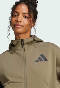 Olijfgroene gewatteerde hoodie met een rits, textuurstof en een zwart grafisch logo op de linkerborst.