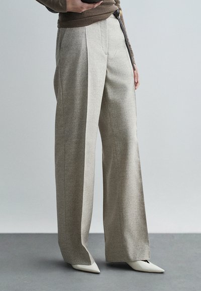 Pantalones de pierna ancha en tejido gris claro con una textura sutil, con pliegues frontales y cintura alta, combinados con zapatos blancos de punta fina.