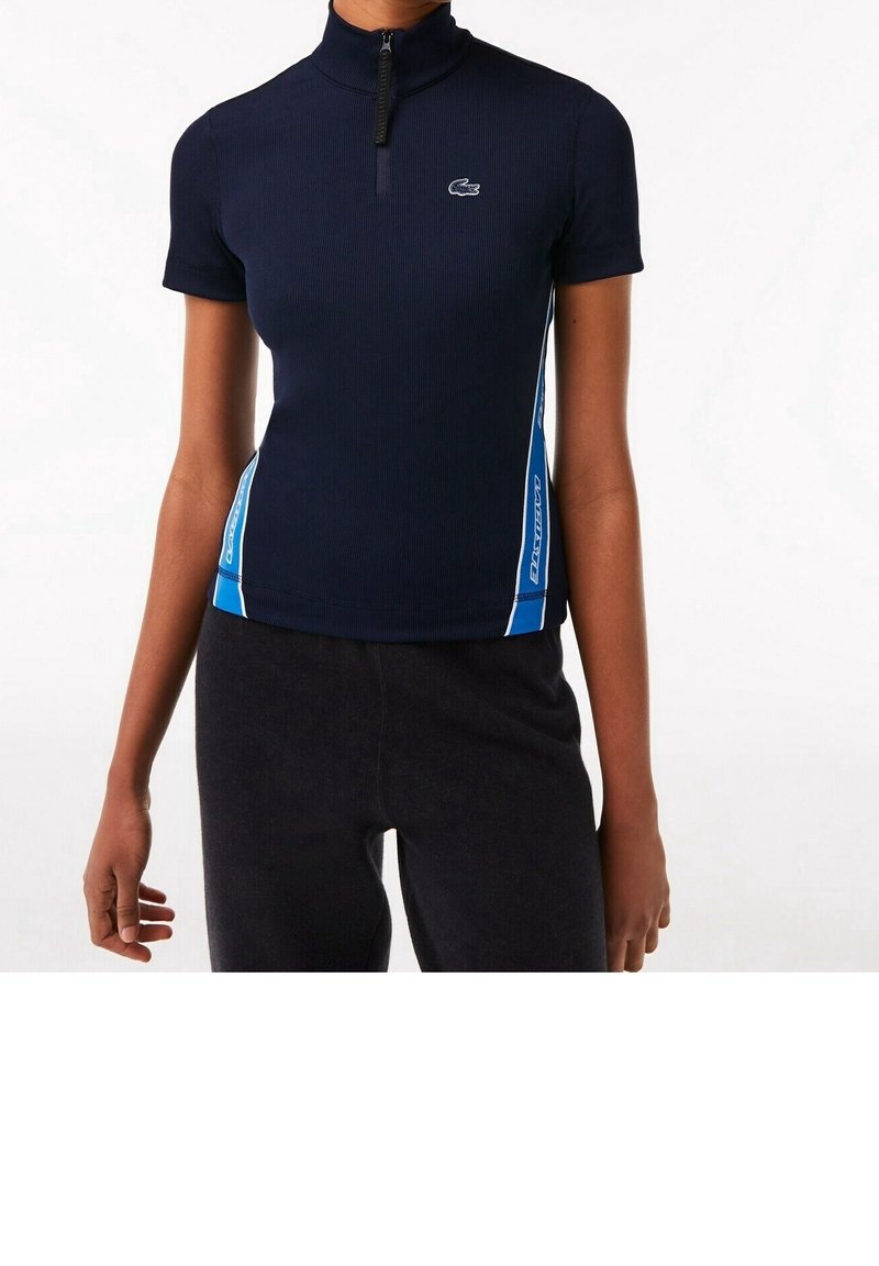 Haut bleu marine à manches courtes avec une demi-fermeture éclair et des bandes latérales bleues et blanches, porté avec un pantalon noir, coupé à mi-torse.