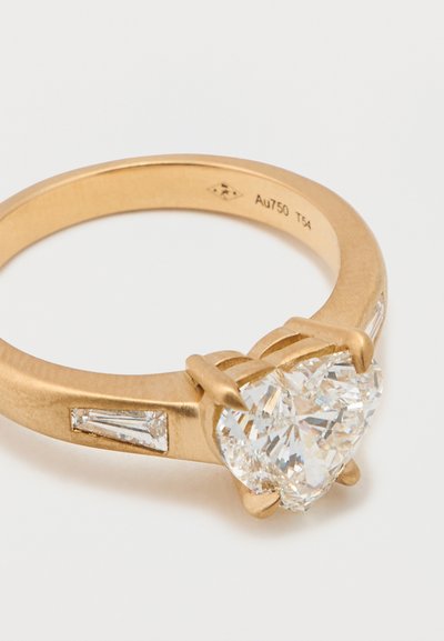 Kullast südamekujuline sõrmus, millel on suur läbipaistev teemant, mille kummalgi küljel on kaks väiksemat ristkülikukujulist teemanti, pael tekstureeritud mattviimistlusega.