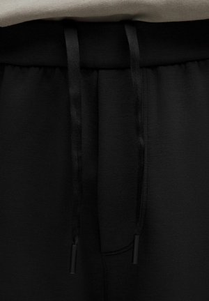 Los joggers negros cuentan con una cinturilla texturizada con cordones negros y aglets negros. Se puede ver un bolsillo en la parte frontal, que muestra una estética limpia.