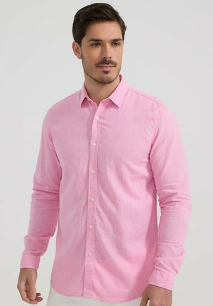 Homme aux cheveux courts et foncés portant une chemise rose à manches longues boutonnée, debout devant un fond clair uni.