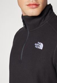 Fekete bolyhos félcipzáras pulóver fehér North Face logóval. Magas nyakkal és sima textúrával rendelkezik, részletgazdag varrásokkal.