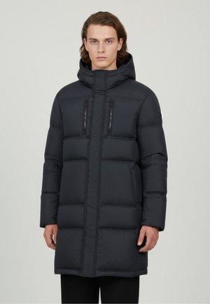Young man wearing a long black padded winter coat with a hood, zipper pockets on the chest, and a high collar, standing against a plain background.
