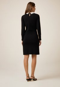 Schwarzes Kleid mit langen Ärmeln, weißem Kragen und Schlüssel-Ausschnitt hinten. Besitzt eine figurbetonte Form und dezente Knopfakzente an den Ärmeln.