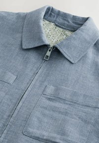 Chaqueta azul de tejido trenzado con cierre de cremallera, dos bolsillos frontales y un forro interior verde con patrón. Tejido texturizado con un ligero brillo.