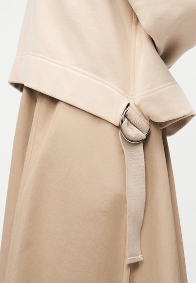 Μπεζ ένδυμα με υφασμάτινη ζώνη περασμένη μέσα από διπλό ασημένιο δαχτυλίδι σε σχήμα D, που δείχνει λεπτομερή ραφή και υφή του υφάσματος.