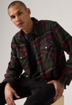 Green and red plaid flannel shirt with two chest pockets, button-up front, and a relaxed fit, layered over a black t-shirt.