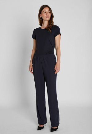 Femme debout, portant un haut noir uni à manches courtes et un pantalon noir ample avec des talons pointus noirs, regardant légèrement vers sa gauche.