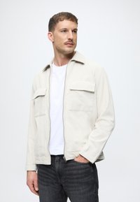 Kremo spalvos kordūrinis švarkas su smailiu apykakle, dviem krūtinės kišenėmis, užtrauktuku ir ilgomis rankovėmis, užsidėtas ant balto marškinėlio ir tamsių džinsų.