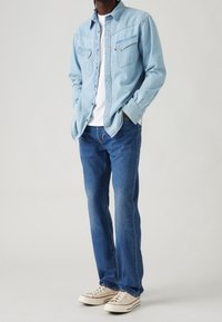 Ljusblå denimskjorta med två bröstfickor, bärs över en vit t-shirt, tillsammans med blå jeans och beige sneakers.