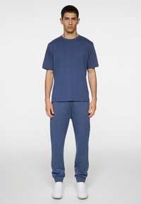 Sininen puuvillainen t-paita pyöreällä kaula-aukolla, yhdistettynä vastaaviin sinisiin collegehousuihin, joissa on nauhavyö ja resorit nilkoissa, käytetty valkoisten lenkkareiden kanssa.