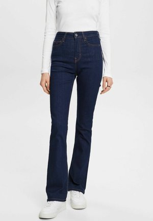 Kvinde iført mørkeblå højtailede vide jeans, hvid langærmet top og hvide sneakers, stående mod en ensfarvet baggrund.