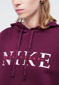 Burgundska kapuca s belim i crvenim 'NIKE' logotipom sportske opreme. Ima kapuljaču sa vezicom i teksturirani materijal. Pogled izbliza na dizajn.