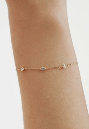 Pulsera delicada de cadena de oro con tres pequeños diamantes redondos espaciados uniformemente, llevada en una muñeca de piel clara sobre un fondo liso.