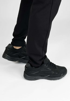 Persoon die zwarte sneakers van mesh en leer draagt met een zwarte broek, staand op een witte vloer.