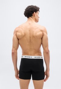 Muskulöser Mann mit lockigem Haar, der ohne Shirt mit dem Rücken zur Kamera steht, trägt schwarze Boxer-Slips mit weißem Bund und der Aufschrift "ONLY&SONS".